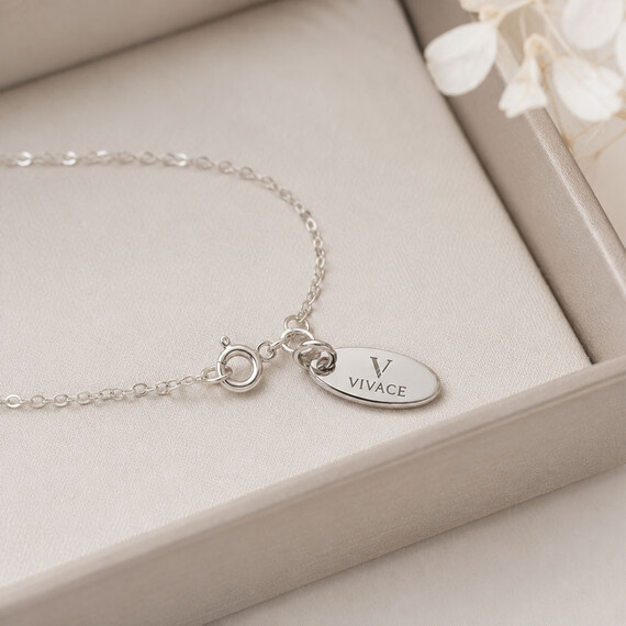 Silver oval jewelry tag engraved with the custom logo and attached near the clasp of a delicate necklace displayed in a presentation box.