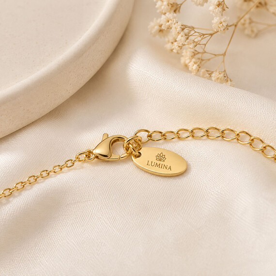 Gold oval jewelry logo tag engraved with custom branding and attached to a fine chain clasp on soft cream satin.