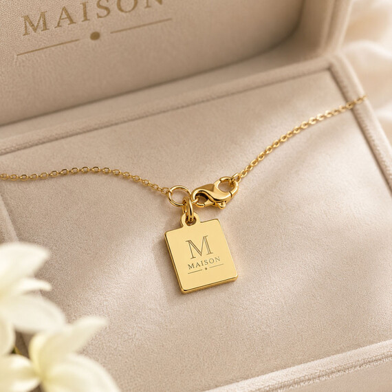 Square gold metal jewelry tag engraved with the custom logo and attached to a necklace clasp inside a premium jewelry box.
