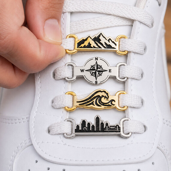 Etiquetas de cordones metálicos personalizadas variadas con diseños de montaña, brújula, ola y horizonte mostradas unidas a los cordones de zapatillas blancas