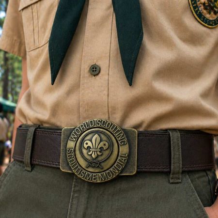 ภาพใกล้ของลูกเสือที่สวมชุดสีกากีและผ้าพันคอสีเข้ม มีหัวเข็มขัดลูกเสือโลกทองเหลืองโบราณที่มีสัญลักษณ์ฟลูร์-เดอ-ลิสอยู่ตรงกลางในบรรยากาศตั้งแคมป์กลางแจ้ง