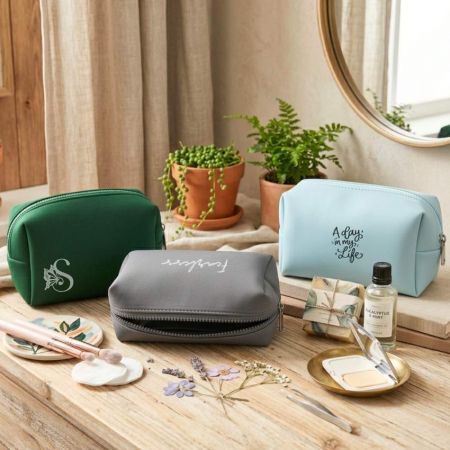 Custom neoprene cosmetic bags in green, gray, and light blue displayed with makeup accessories on a wooden table