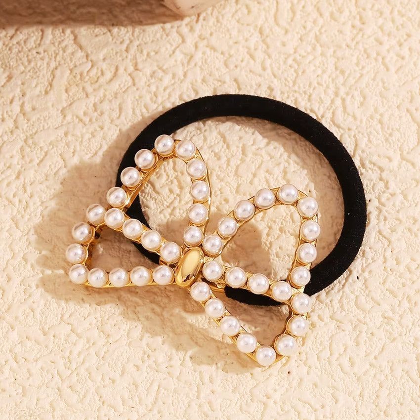 A close-up of the stainless steel butterfly pearl charm hair tie in a gold finish, showing the intricate pearl details on the butterfly charm attached to a black elastic band.