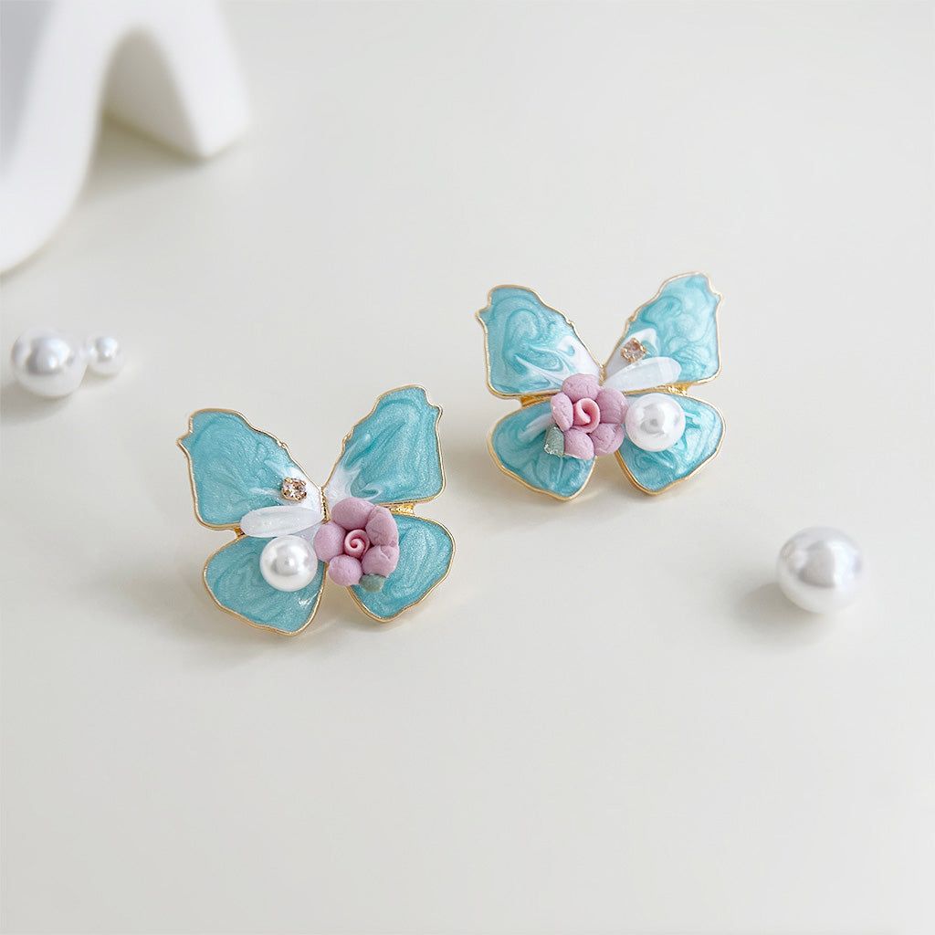 Un par de pendientes de mariposa de esmalte azul chapados en oro con delicados detalles de perlas y flores sobre un fondo blanco limpio.