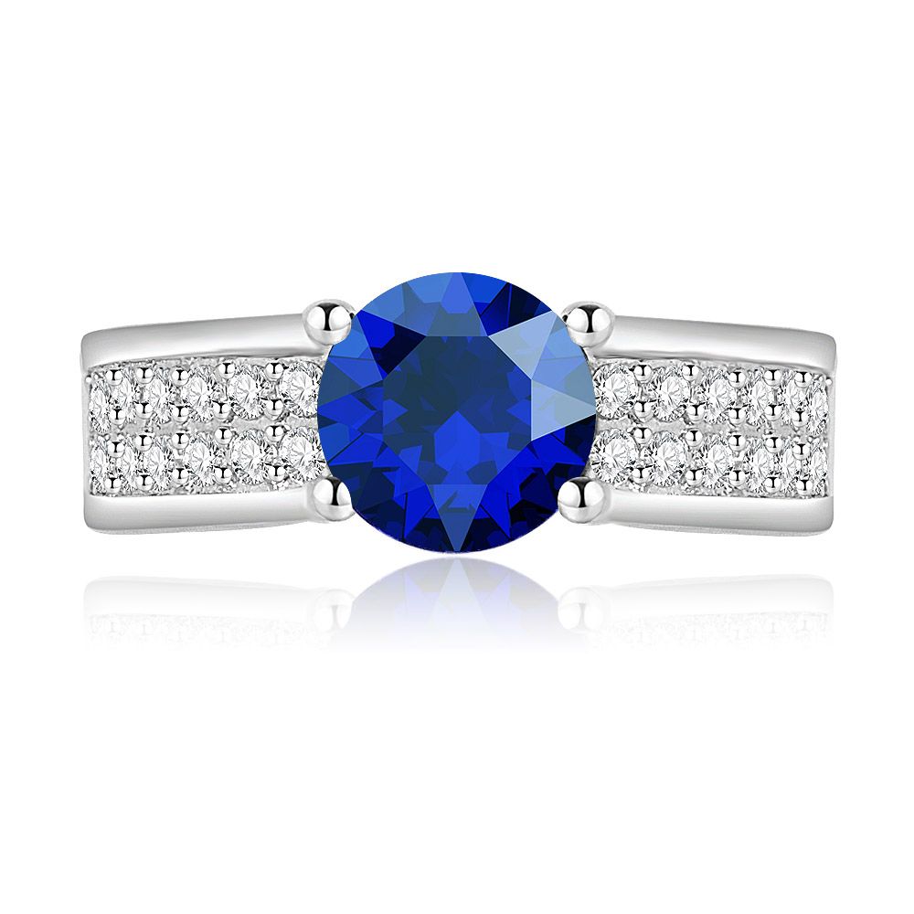 Close-up van de ronde saffier- en zirconia-ring, met de prominente blauwe gecreëerde saffiersteen omringd door een dubbele rij sprankelende zirconia op een rhodium-geplateerde band.