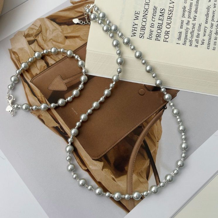 Un sofisticado conjunto de collar y pulsera de perlas grises de plata esterlina 925, elegantemente exhibido junto a un clásico bolso de cuero marrón.