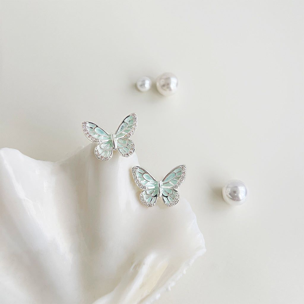 A pair of wholesale silver-plated butterfly stud earrings with mint-green enamel and clear rhinestones, elegantly displayed on a white seashell.