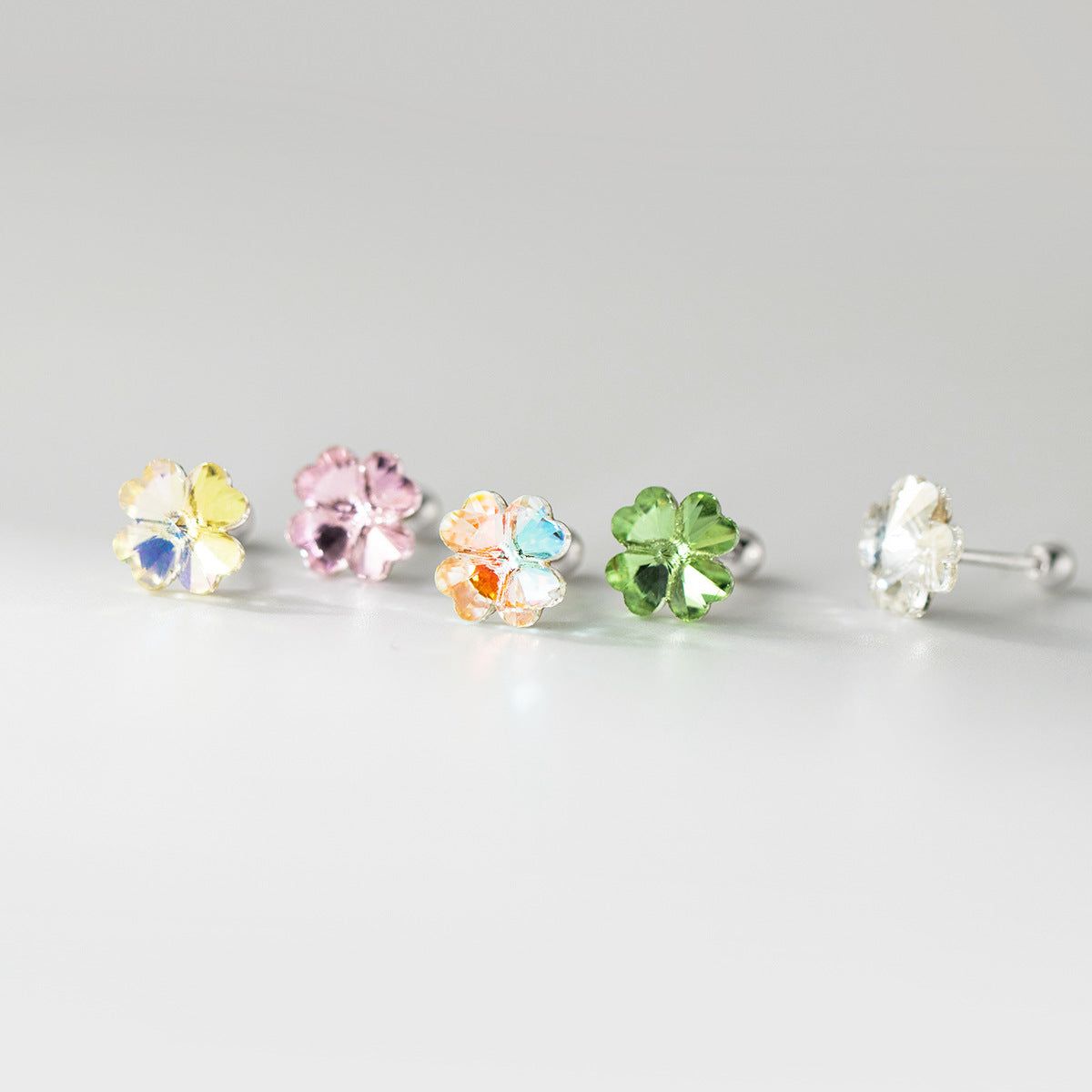 Uma coleção vibrante de cinco pares de brincos de trevo de quatro folhas em prata esterlina 925 com pedras preciosas de cristal coloridas, exibidos em um fundo branco limpo.