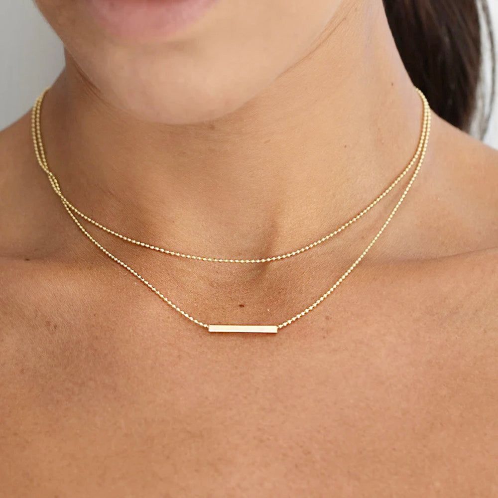 Um colar estiloso de dupla camada em aço inoxidável com acabamento dourado, apresentando um pingente de barra elegante em uma delicada corrente de bolinhas.