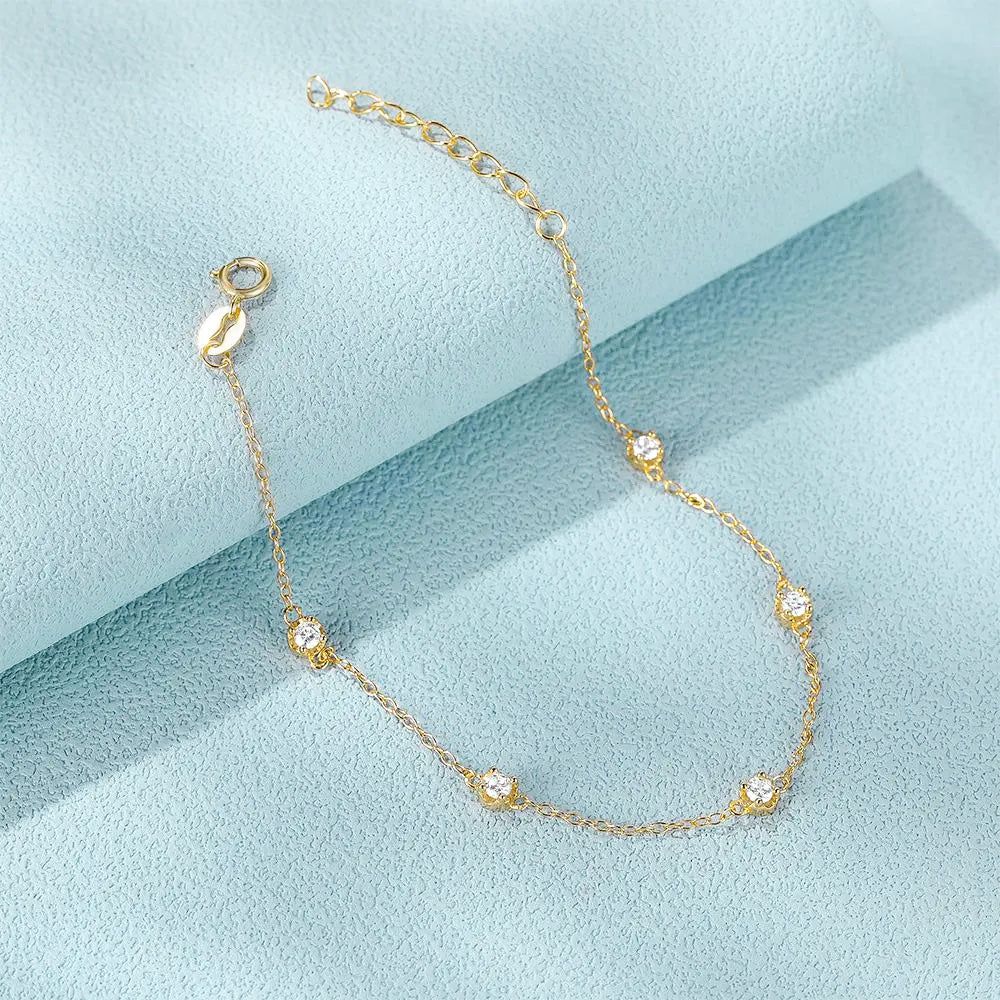 Una delicada pulsera de cadena de plata esterlina 925 en acabado dorado, con cinco piedras de circonita brillantes, exhibida sobre un fondo azul claro.