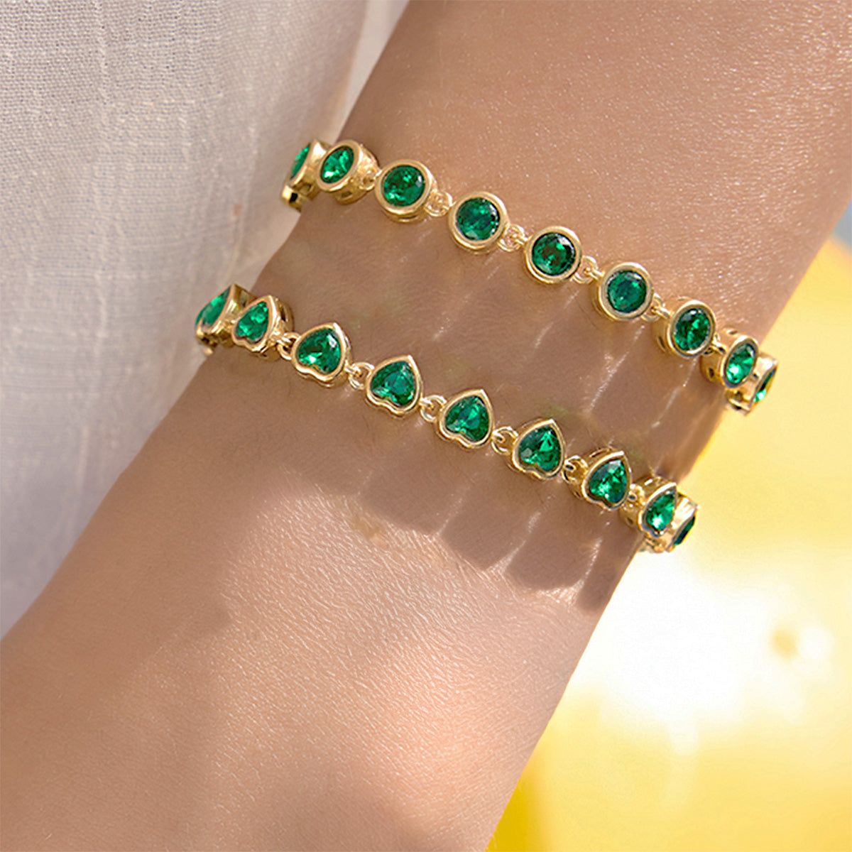 En kvinnas handled prydd med setet av armband i rostfritt stål med gröna hjärtan, som framhäver de briljanta gröna hjärtstenarna och den eleganta guldtonade finishen.