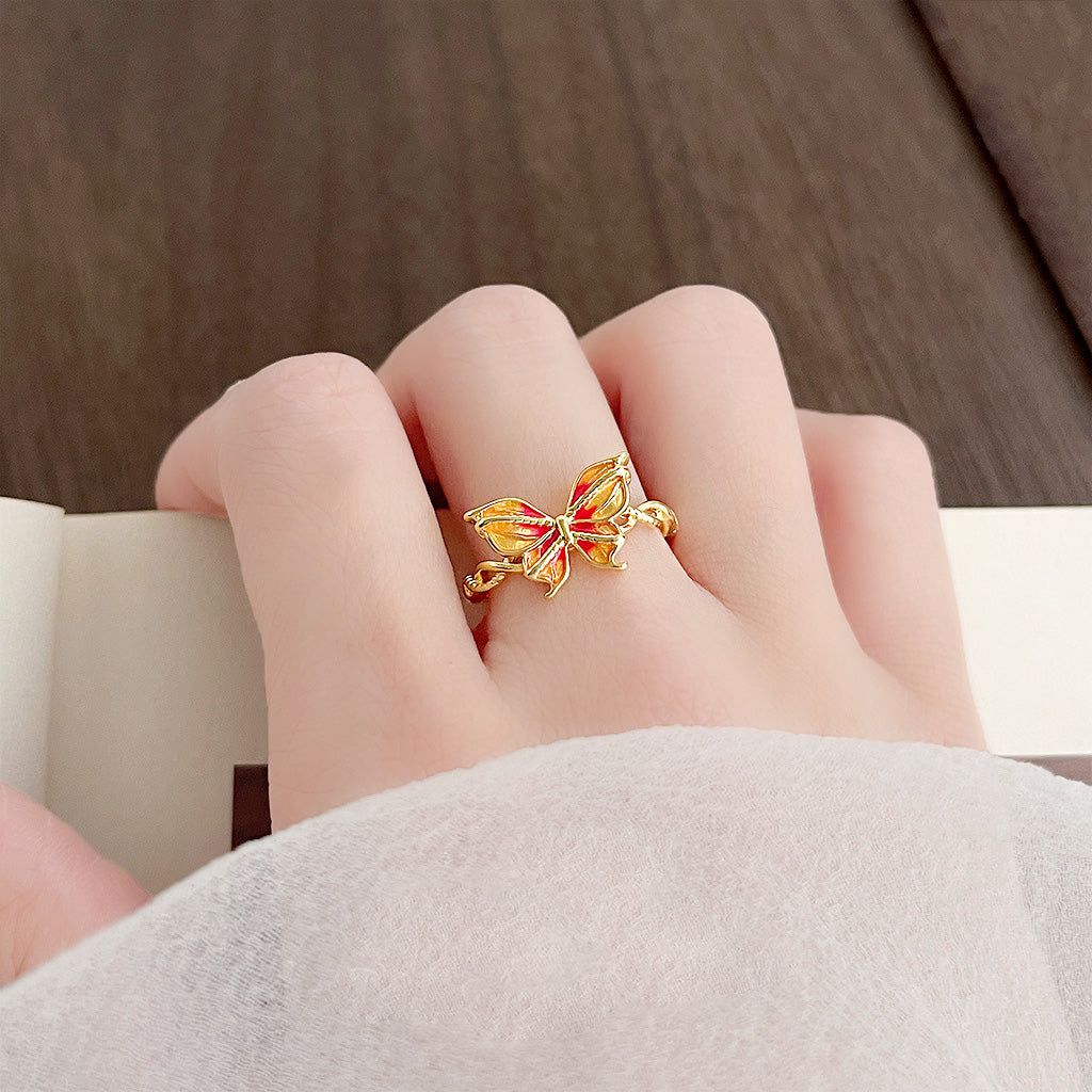 A close-up of a gold-plated adjustable ring featuring a delicate butterfly with gradient orange and red enamel wings.