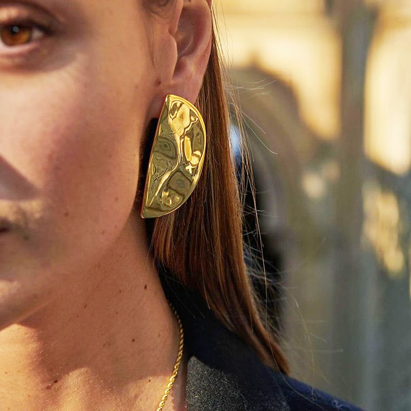 Una vista en primer plano de una mujer usando un pendiente de media luna de acero inoxidable dorado con un acabado martillado brillante, mostrando su textura y diseño.