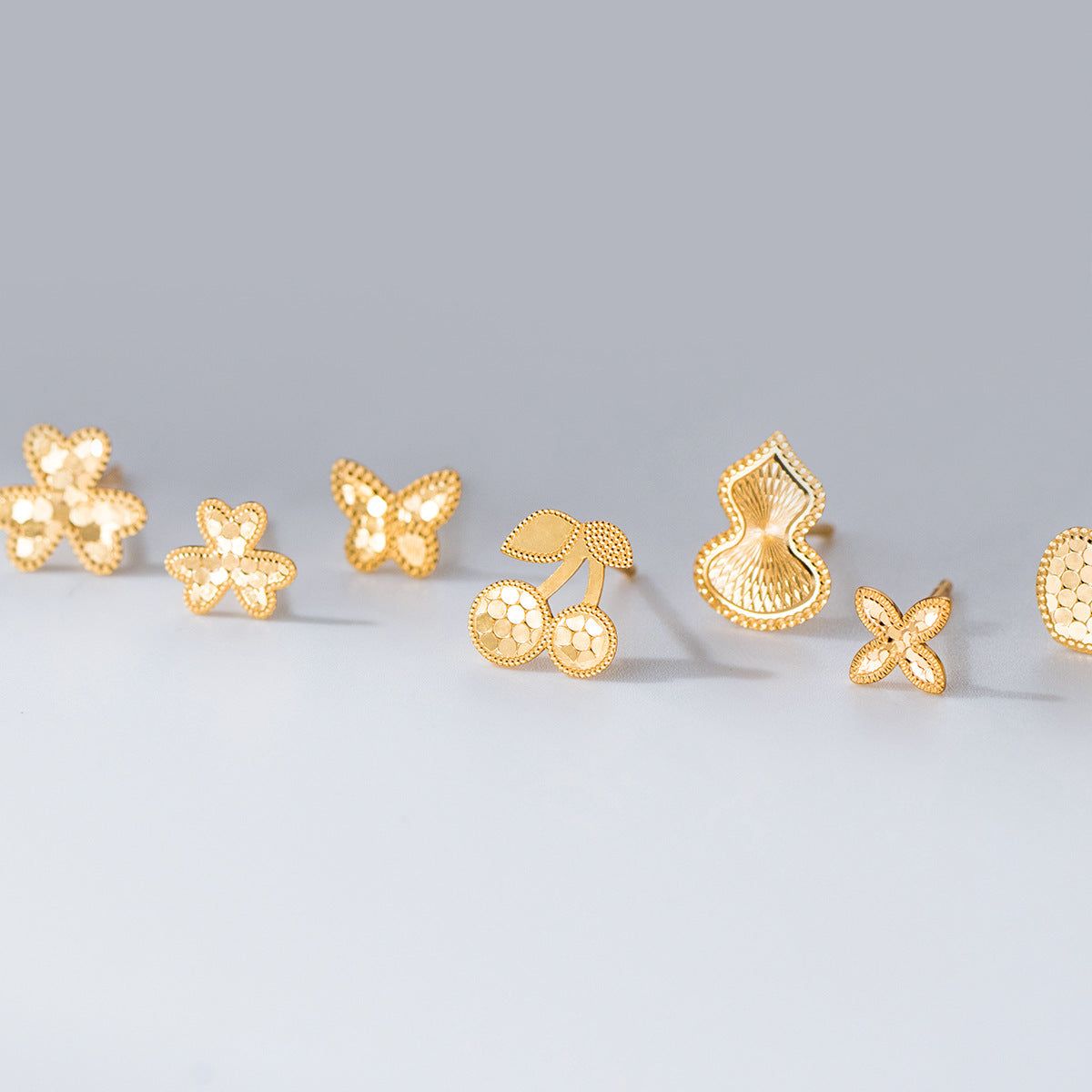 Eine Kollektion von fünf Paaren von Großhandels-Ohrsteckern aus 925 Sterling Silber mit Lasergravur in Gold, mit Designs von Blumen, Schmetterlingen, Herzen, Kirschen und Gourds, auf einem weißen Hintergrund angeordnet.