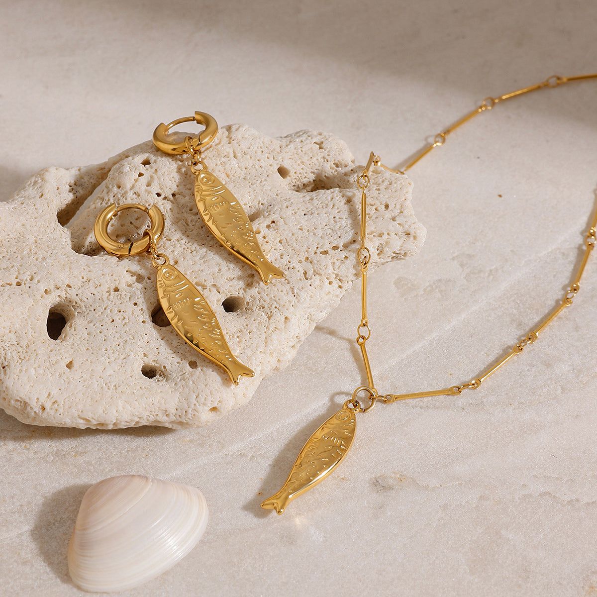 Un set di gioielli a forma di pesce d'oro straordinario, che include una collana e orecchini, elegantemente esposto su una pietra beige con una conchiglia, perfetto per stili a tema spiaggia.