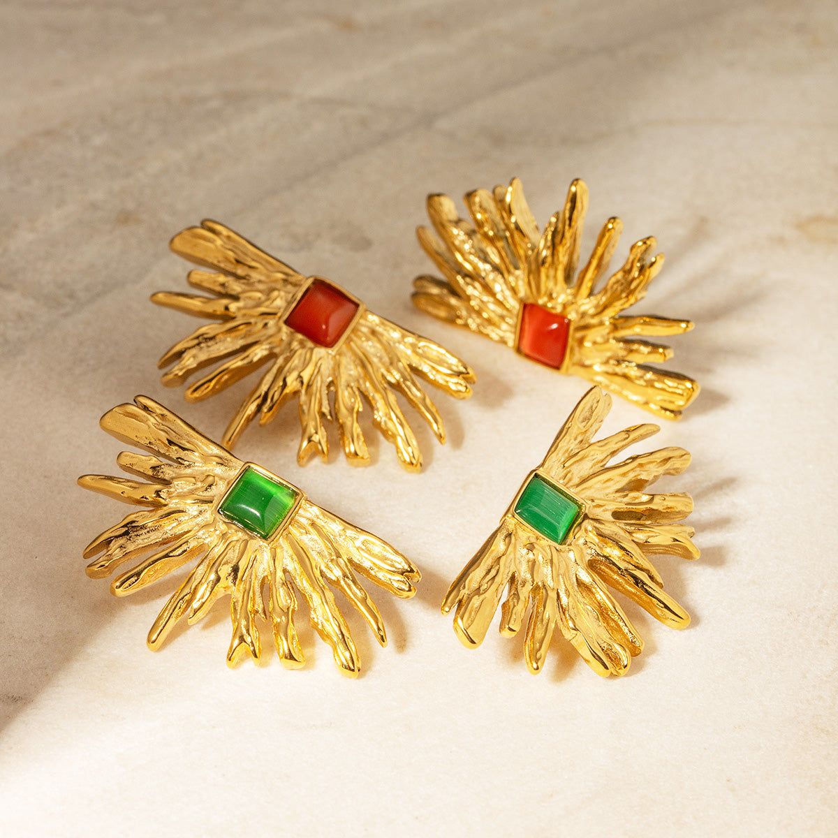 Een collectie van roestvrijstalen, goudgeplateerde drama waaier-vormige stud oorbellen, met zowel de groene als rode edelsteenopties op een witte achtergrond.