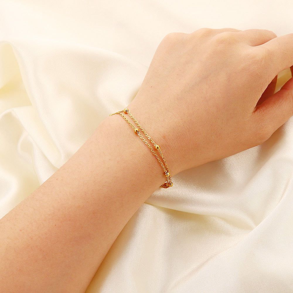Een close-up van de roestvrijstalen dubbele laag kralenarmband met een hartbedel, gedragen om een pols, die de elegante gouden afwerking toont.