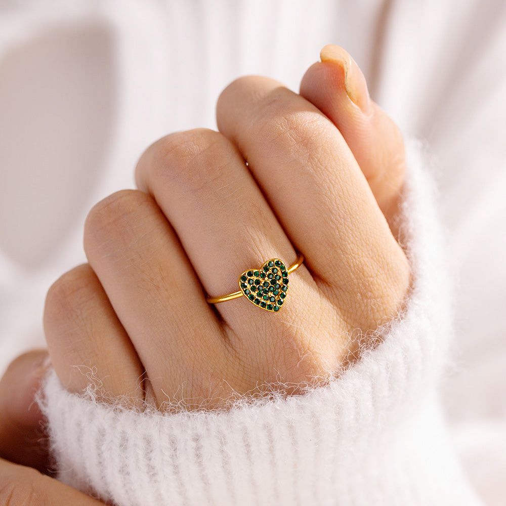 Eine Nahaufnahme eines vergoldeten Edelstahlrings mit einem grünen Zirkonherz, das an einem Finger getragen wird, um sein elegantes Design zu zeigen.