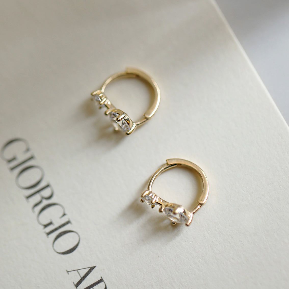 Un paio di orecchini a cerchio mini huggies in cristallo in argento sterling 925 con finitura dorata e gemme chiare, esposti su uno sfondo bianco.