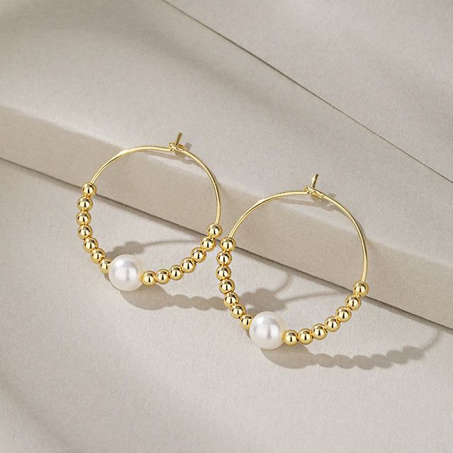 Un paio di eleganti orecchini a cerchio in argento sterling 925 con finitura dorata, adornati con una perla centrale e delicate perline dorate.