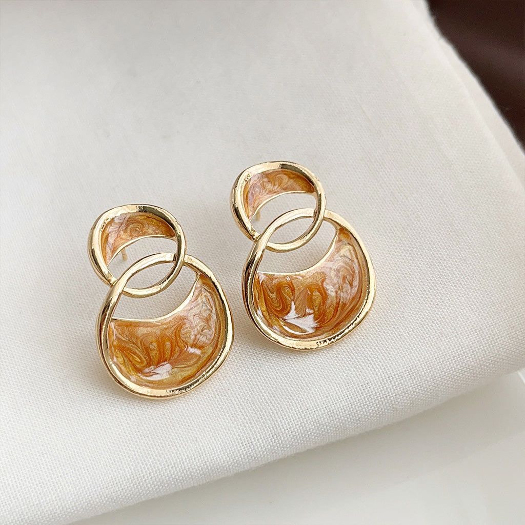 Gros plan sur les boucles d'oreilles en clou plaquées or 14k, mettant en avant le design distinctif à double boucle avec un motif d'émail marbré camel et blanc.