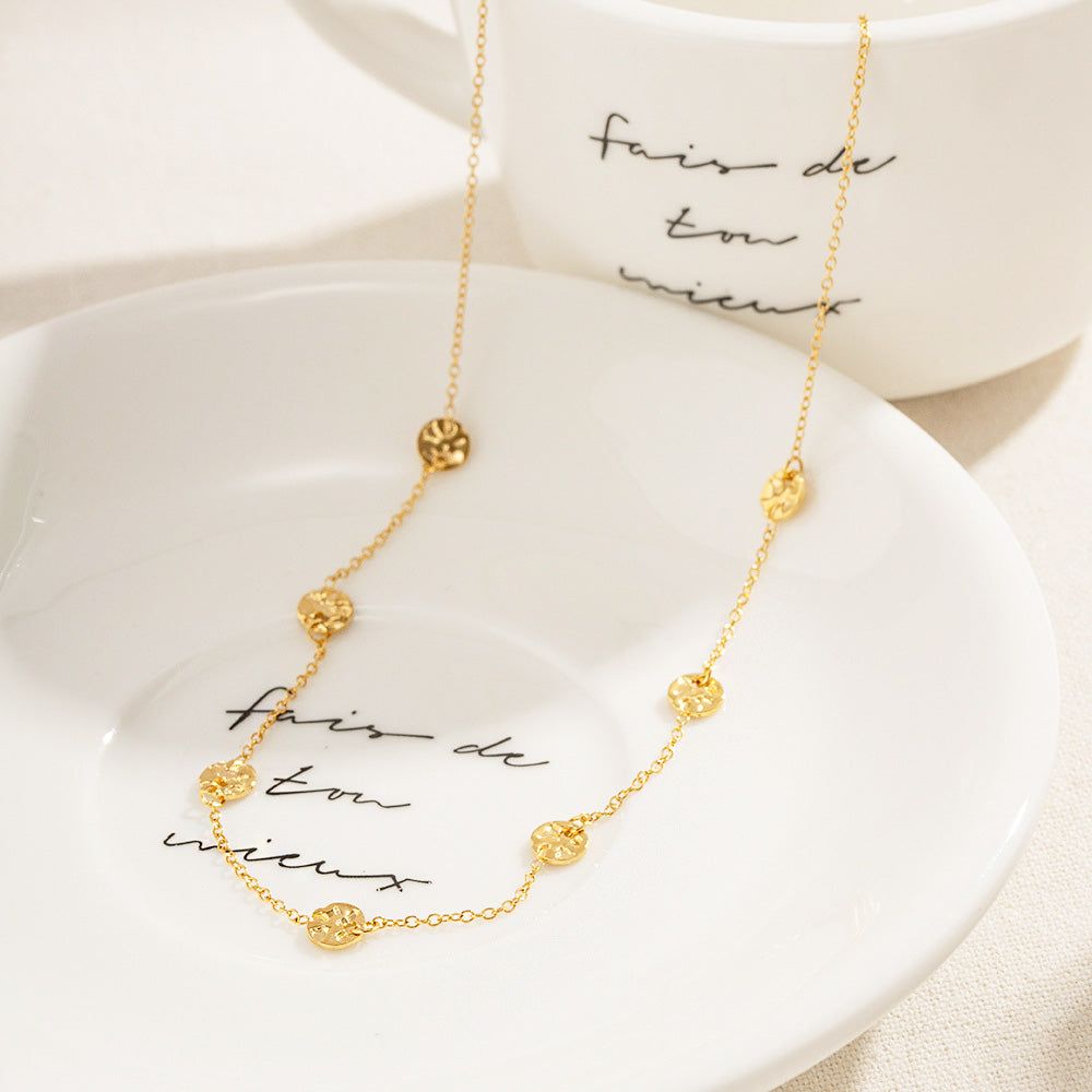 Ett set av guldiga rostfria stålchokerhalsband med små hamrade myntskivor, elegant uppvisade på en dekorativ tallrik.
