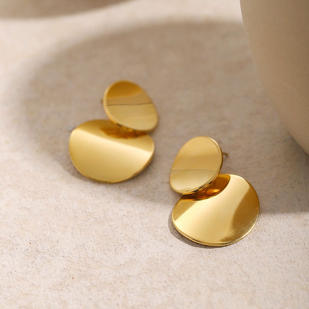 Een paar geometrische drop oorbellen van roestvrij staal met een gouden afwerking, met een elegante gelamineerde en overlappende schijfontwerp, op een schone witte achtergrond.