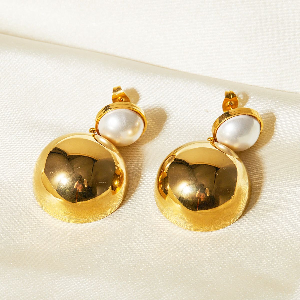 Un par de pendientes colgantes de acero inoxidable chapados en oro con acentos de perla incrustados y un diseño de cúpula hemisférica brillante, mostrados contra un fondo blanco.