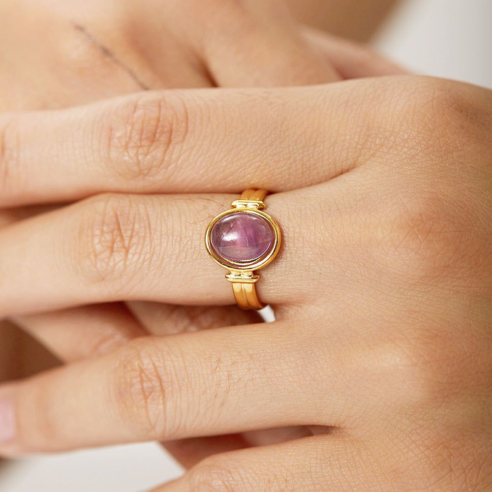 Een close-up van een goudafgewerkte roestvrijstalen open ring met een gepolijste paarse amethist cabochon edelsteen, op een schone witte achtergrond.