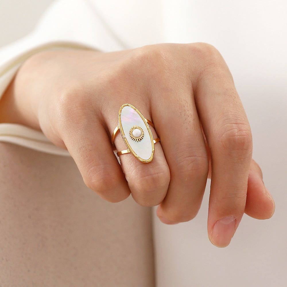 Gros plan d'une bague en acier inoxydable plaqué or du set, avec un accent en nacre blanche ovale et une perle centrale, portée sur un doigt.