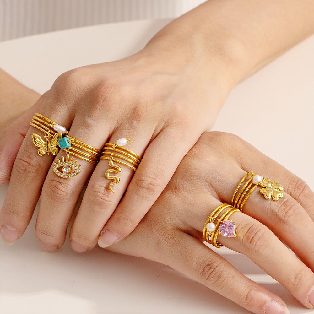 Uma coleção de cinco anéis de aço inoxidável banhados a ouro 18K com vários encantos, como uma borboleta, olho grego, cobra e pedras preciosas, exibidos em um fundo neutro.