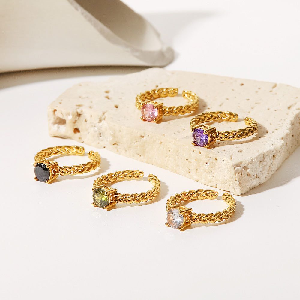 Um conjunto de cinco anéis abertos de aço inoxidável banhados a ouro, cada um com uma pedra de zircônia cúbica de cor diferente, exibidos em um fundo branco limpo.