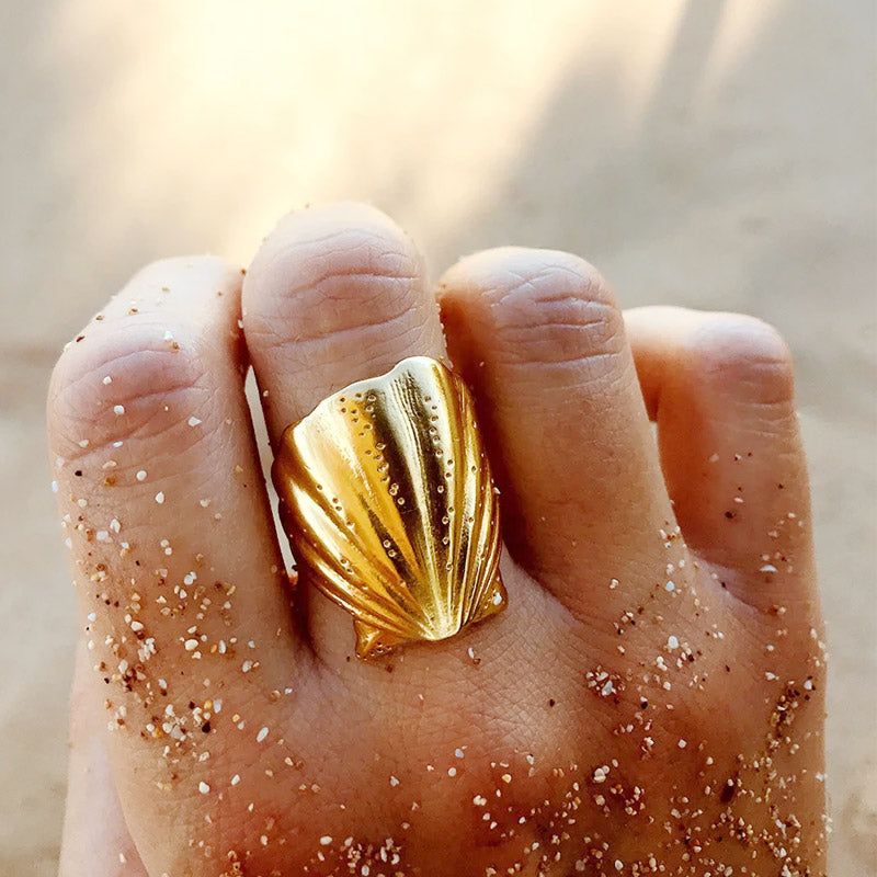 La mano di un modello che indossa un anello aperto a forma di conchiglia in acciaio inossidabile, resistente all'ossidazione, poggiato su una superficie sabbiosa.