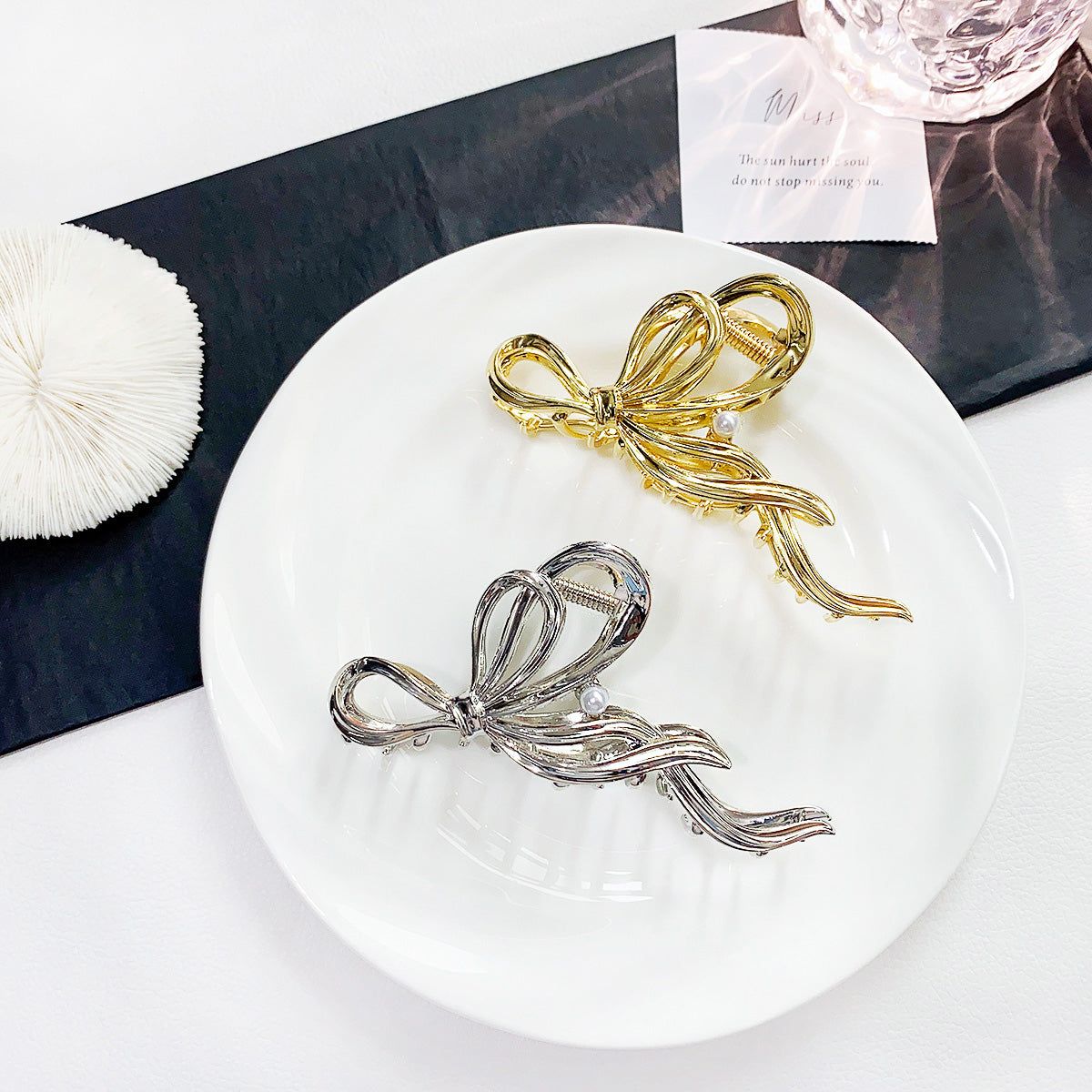 A pair of elegant metal bow-shaped hair claw clips, one in a polished gold finish and the other in a sleek silver finish, displayed on a white surface.