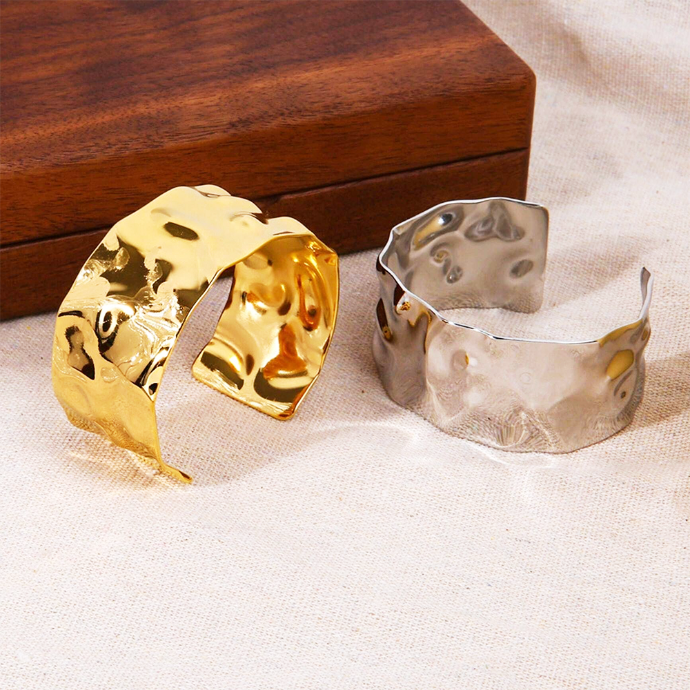 Ett set av breda armband i rostfritt stål med en hamrad textur, där ett har en polerad guldfinish och ett har en elegant silverfinish.