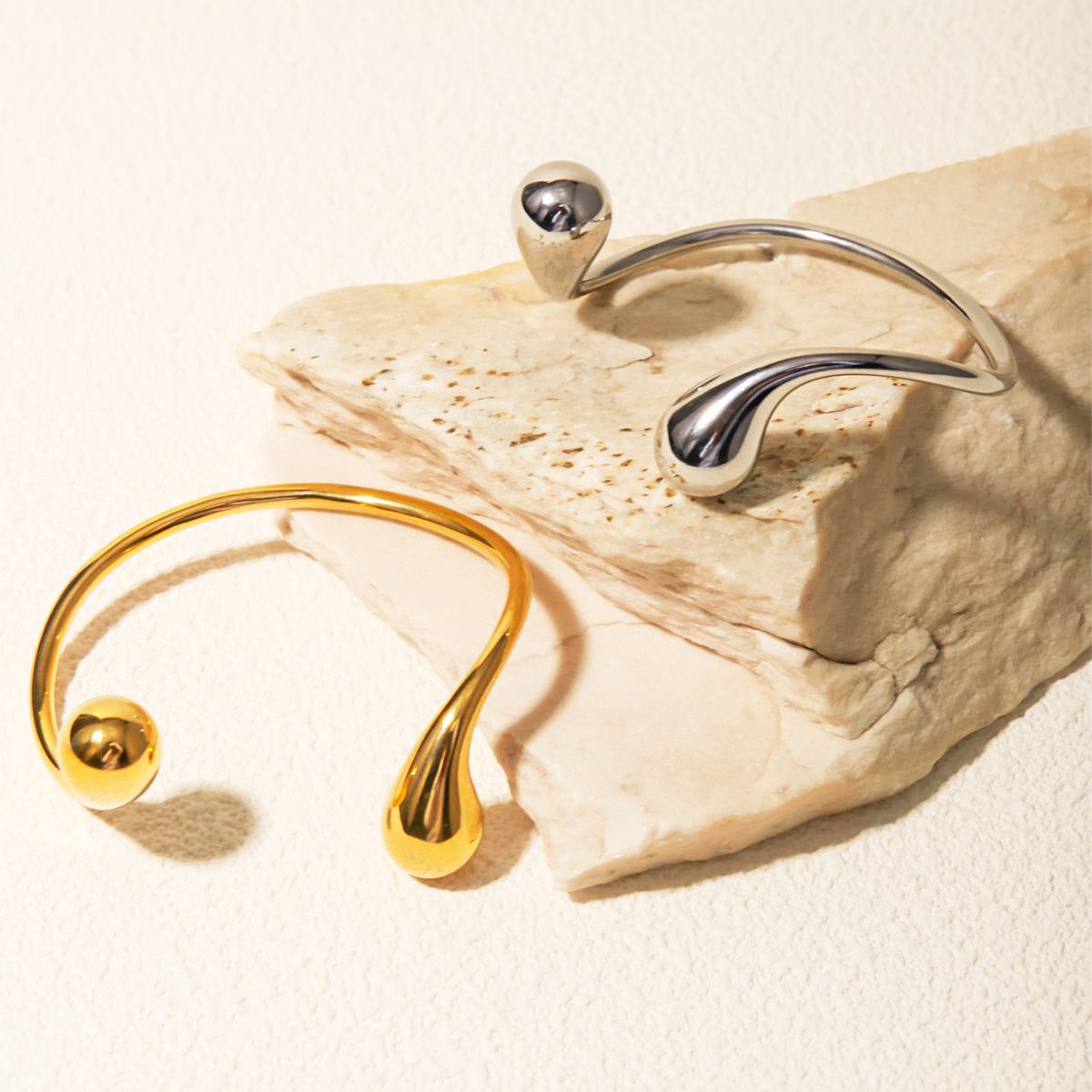 Een set van twee roestvrijstalen cuff bangles, één goud en één zilver, met een modern asymmetrisch druppelontwerp, weergegeven op een gestructureerd stenen oppervlak.