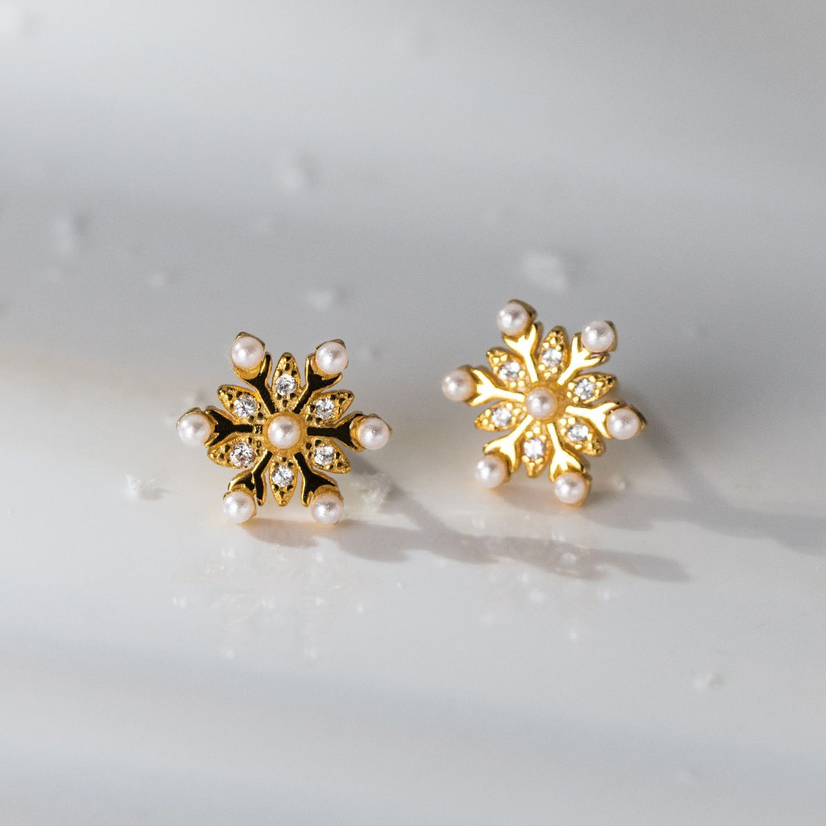 Een paar gouden sneeuwvlok stud oorbellen gemaakt van 925 sterling zilver, met een centrale parel en sprankelende cubic zirconia accenten.