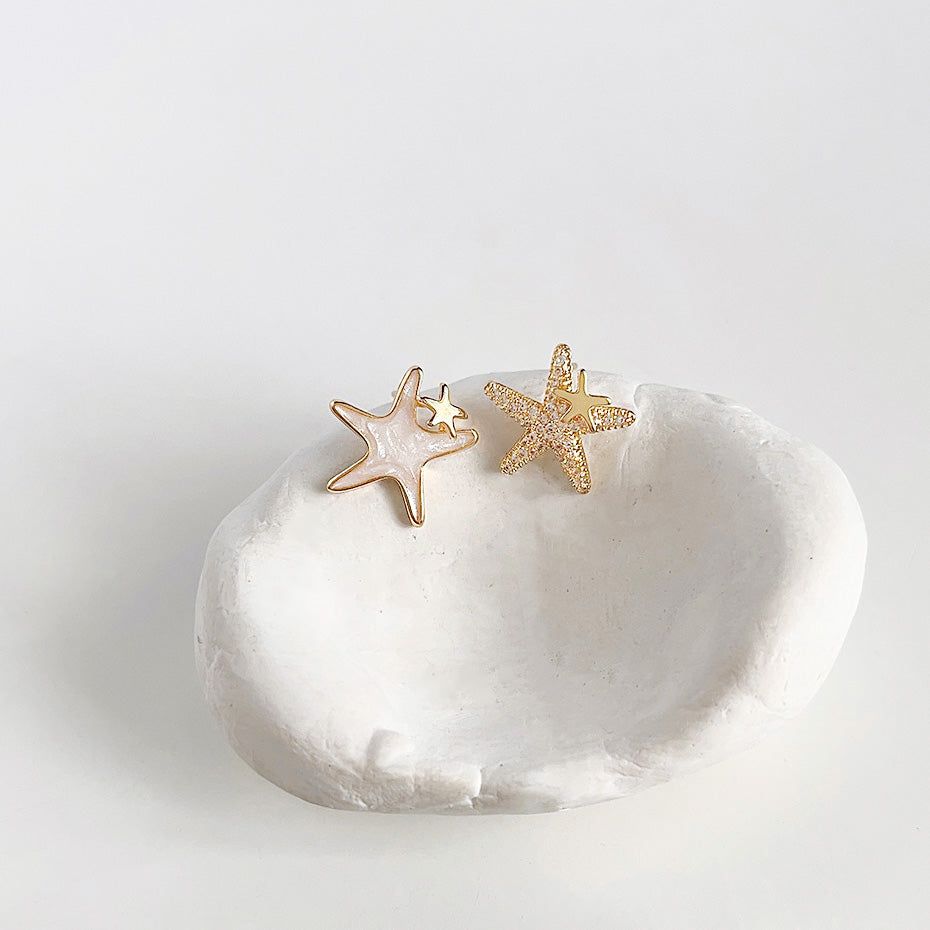 Um par de brincos assimétricos de estrela banhados a ouro 14k de um conjunto de cinco, um com acabamento em ouro texturizado e o outro com esmalte branco, exibidos sobre uma pedra decorativa.