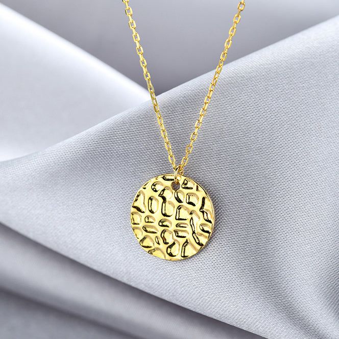 Primer plano de un collar de plata esterlina 925, chapado en oro, con un colgante de disco de moneda geométrico con una textura martillada distintiva.