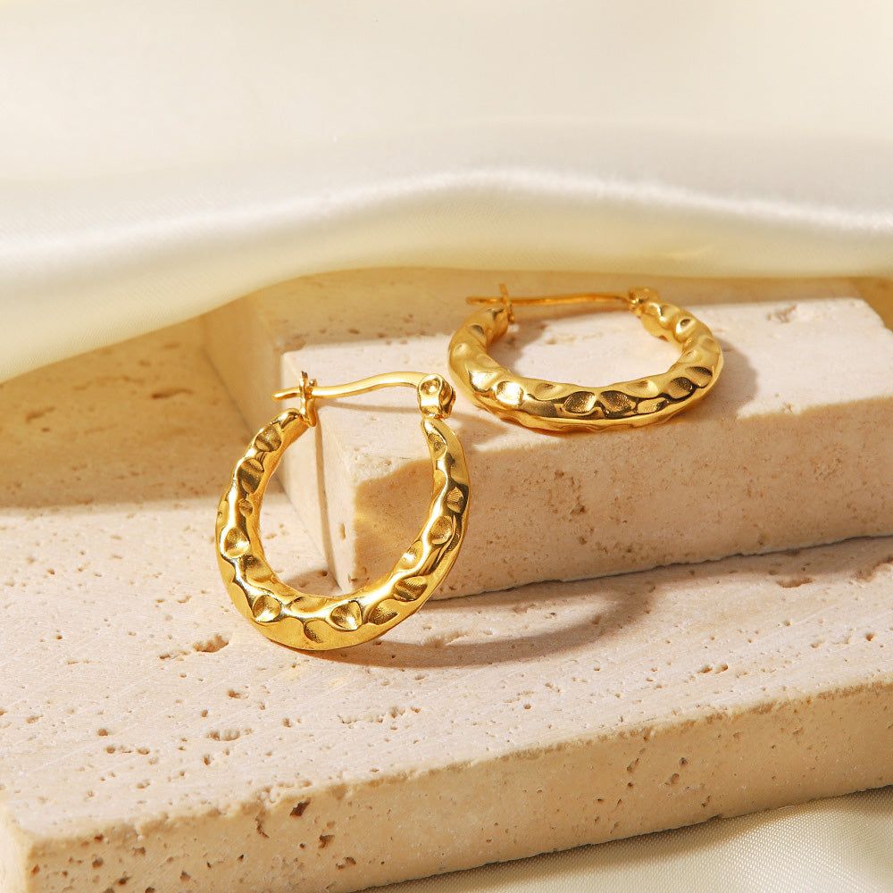 Un par de pendientes de aro dorados en acero inoxidable con una chic textura martillada, exhibidos sobre un fondo blanco limpio.