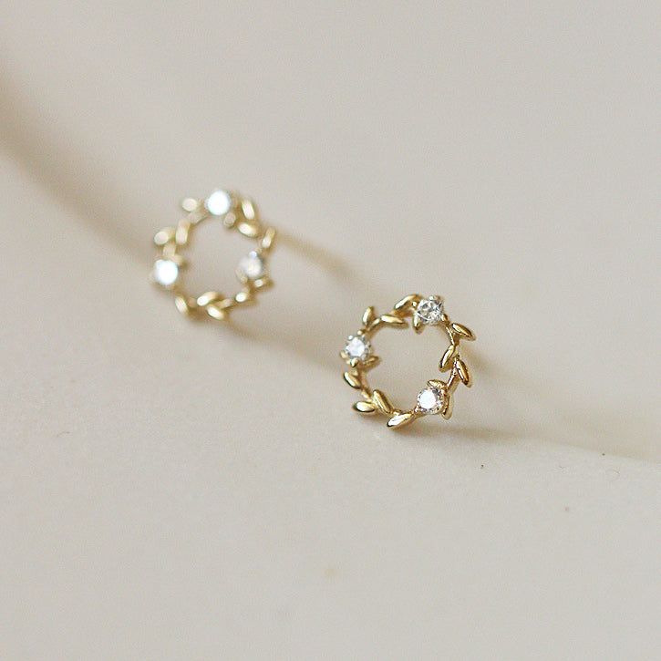 Un primo piano degli orecchini a bottone a forma di corona d'oro piccoli in argento sterling 925, evidenziando il design intricati delle foglie e le pietre bianche scintillanti.