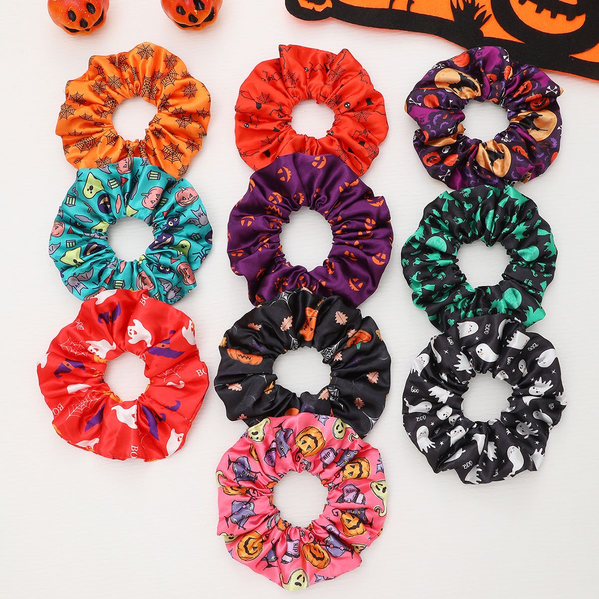 A vibrant collection of Halloween-themed hair scrunchies in various colors and patterns, including pumpkins, ghosts, and spiders, offered by JR Fashion Accessories.