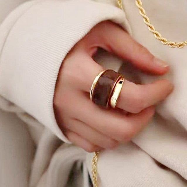 A close-up of a hand wearing two brass cuff rings with a chunky, dual-layered chocolate enamel design, part of a stylish set of five.