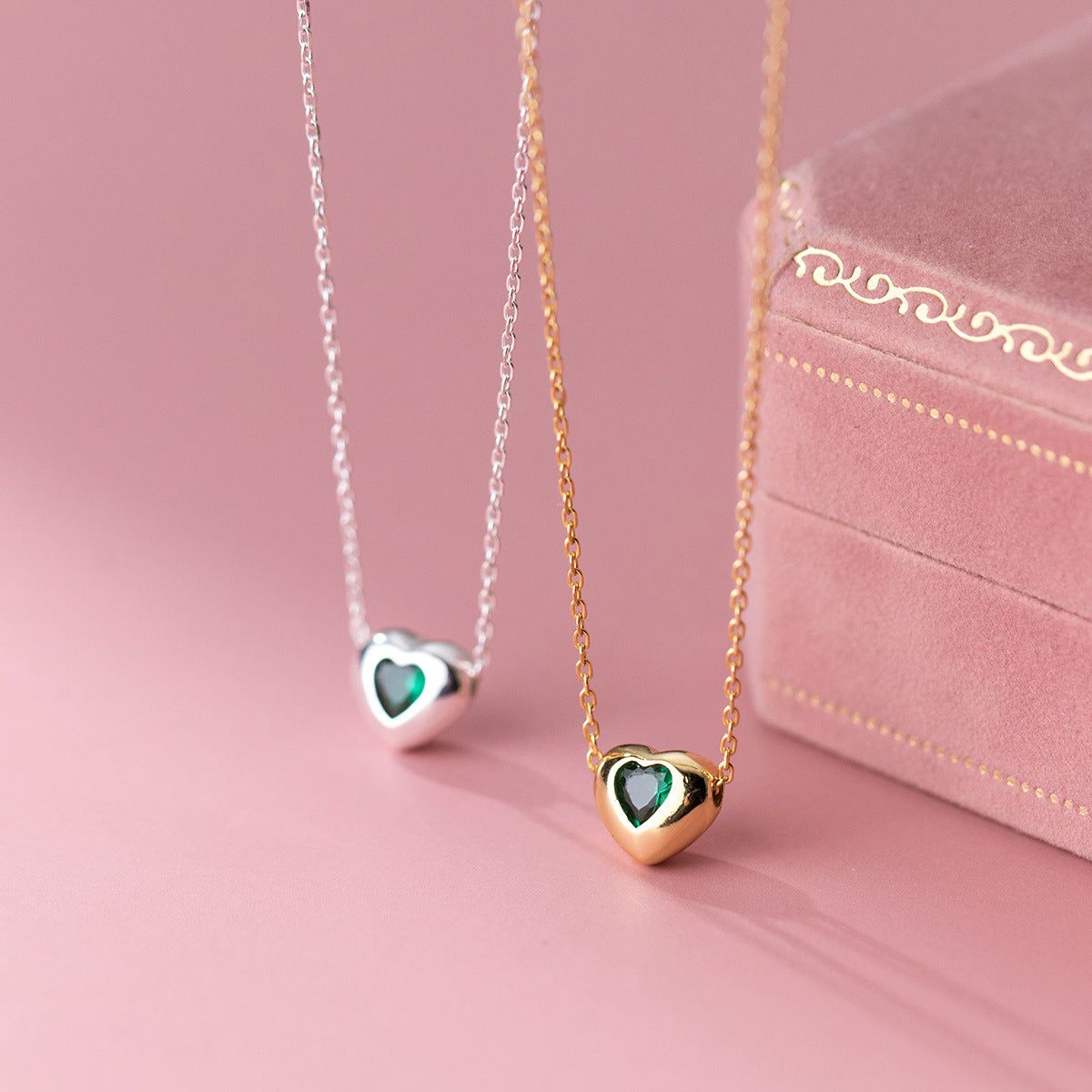 A set of 925 sterling silver heart-shaped necklaces in gold and silver finishes, featuring sparkling green zircon stones, displayed with a pink velvet jewelry box.