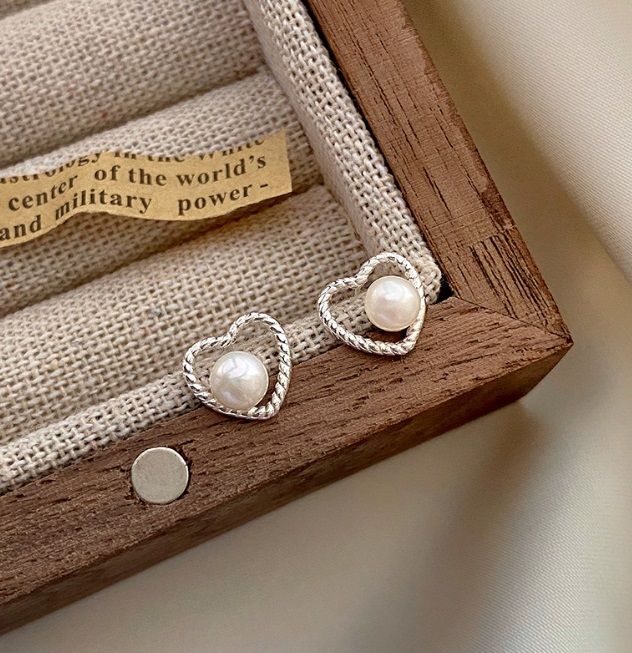Un paio di orecchini a forma di cuore in argento sterling 925 con una perla centrale, elegantemente esposti all'interno di una scatola di legno per gioielli.