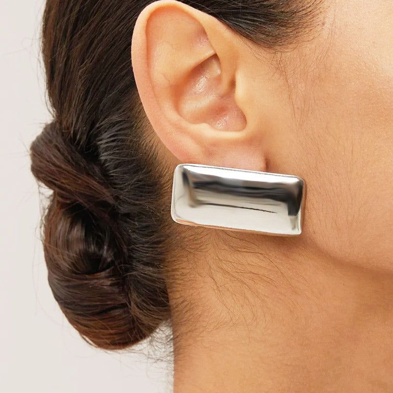 A close-up shot of a woman wearing a large, chunky rectangular stud earring in a polished silver finish, highlighting its modern geometric design.