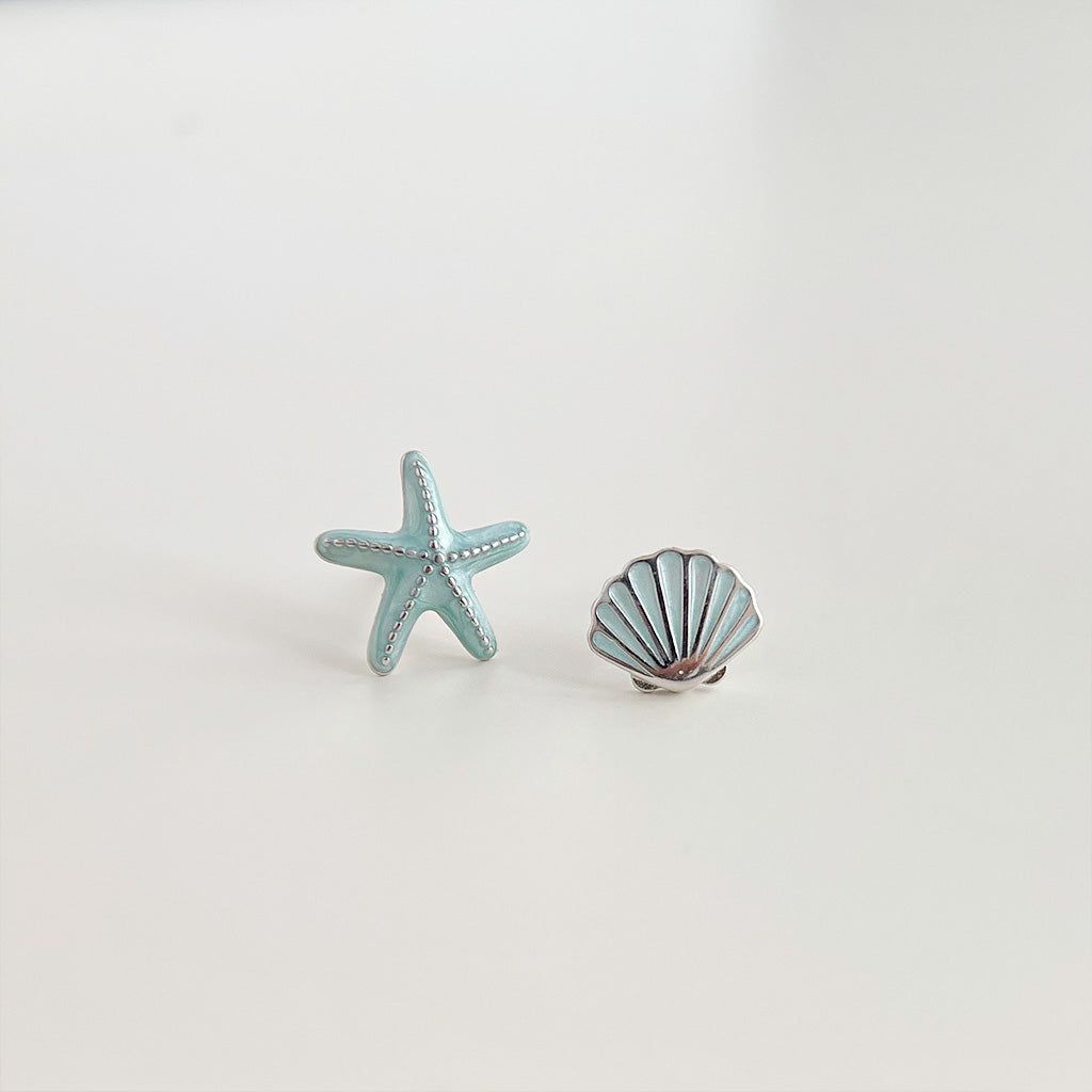 A pair of wholesale silver-plated enamel stud earrings featuring a light blue starfish and a silver seashell on a white background.