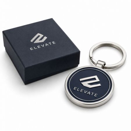 Llavero promocional de metal plateado redondo relleno con esmalte suave azul oscuro corporativo al lado de una caja de regalo premium.