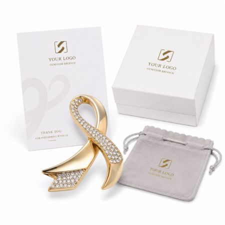 Custom OEM ODM gold awareness ribbon brooch encrusted with rhinestones beside branded gift packaging.
