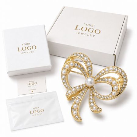 Customizable gold bow brooch set with pearls and rhinestones, shown with wholesale shipping boxes.